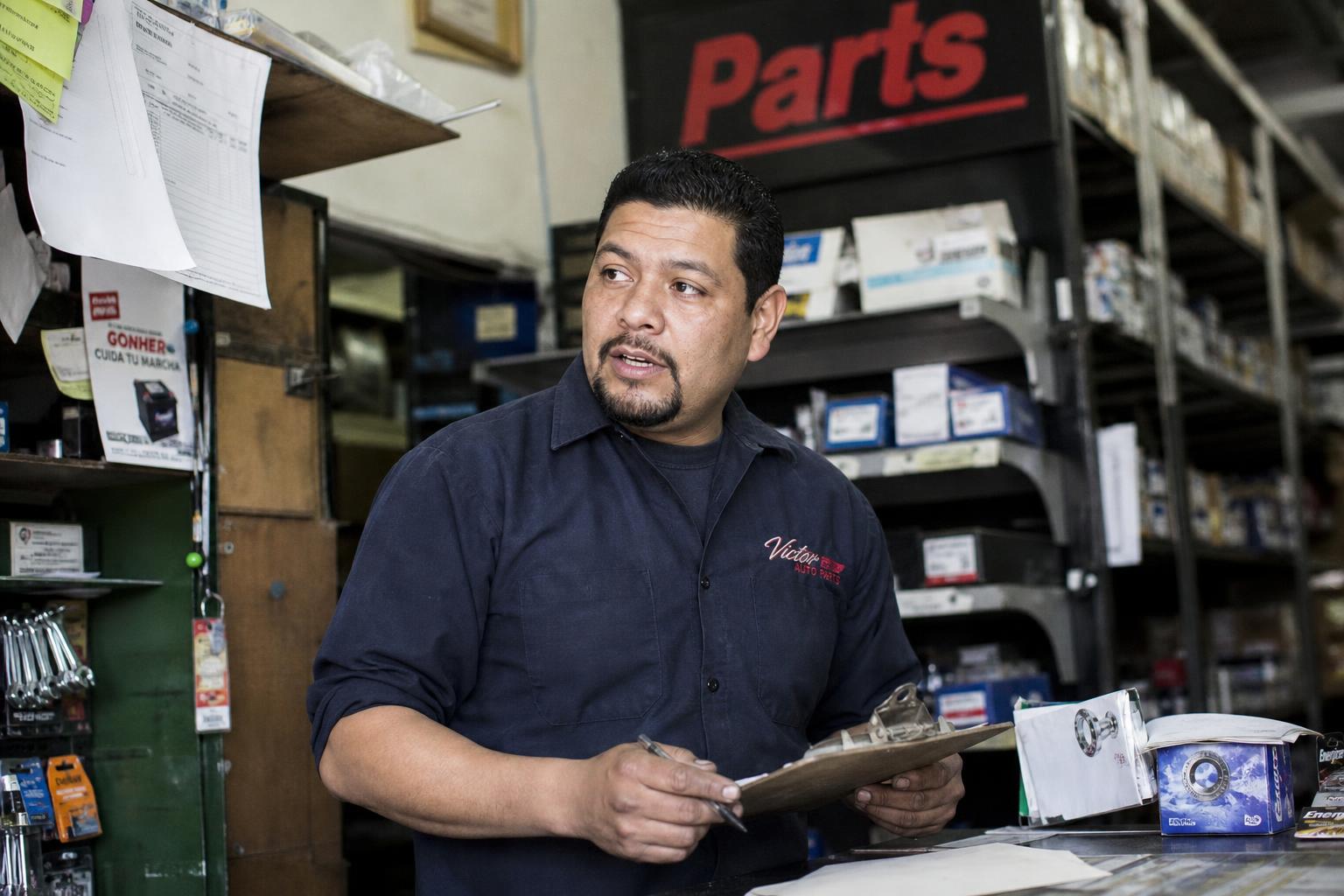 Portrait of Alex Rivera, OEM Parts Advisory Team Lead, standing at the parts counter in a red and gray work shirt, shelves of labeled parts behind him.