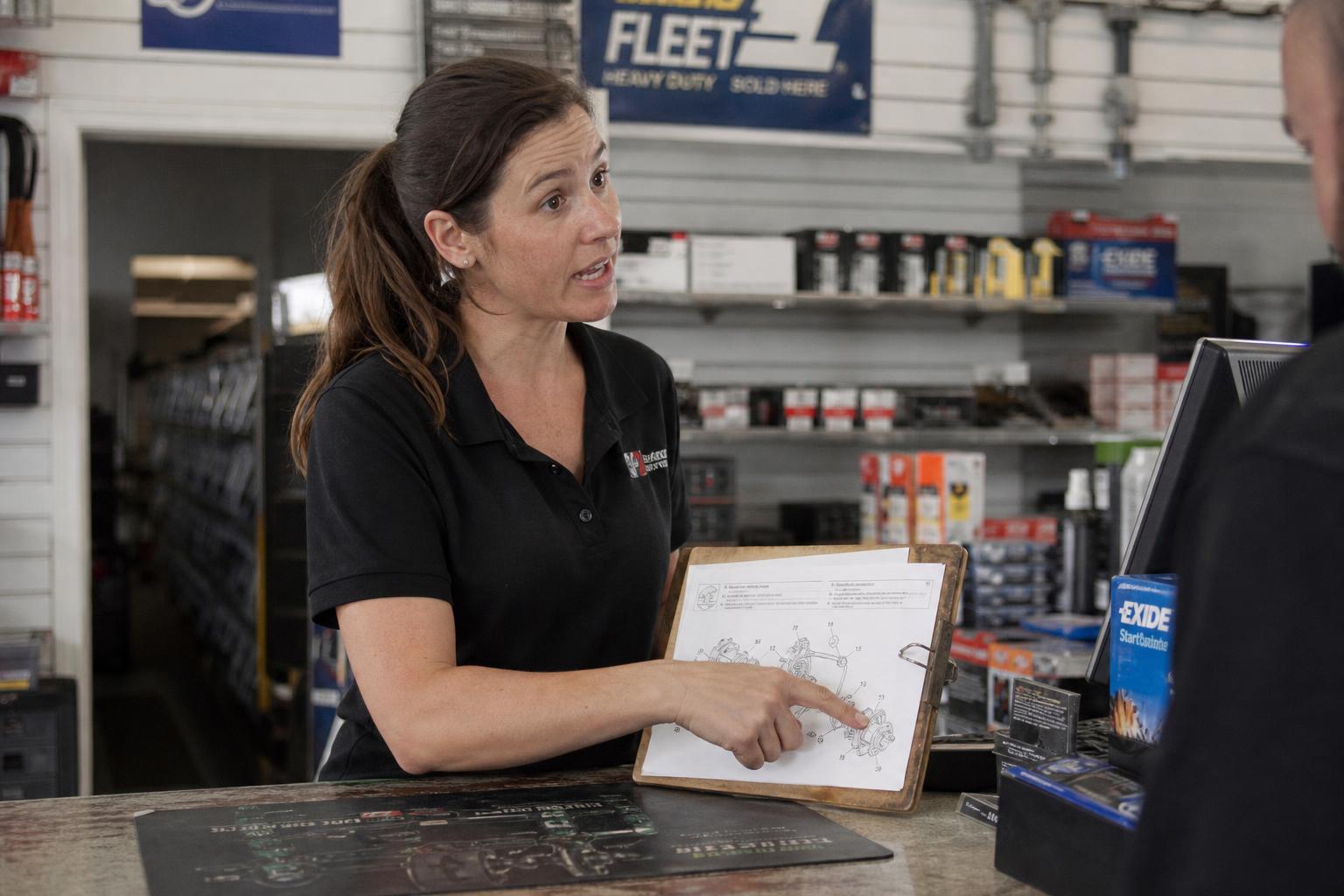 Portrait of Sarah Johnson, Vehicle Parts Consultant, at the service counter in a charcoal work polo, order terminal and fitment catalogs visible on the desk.